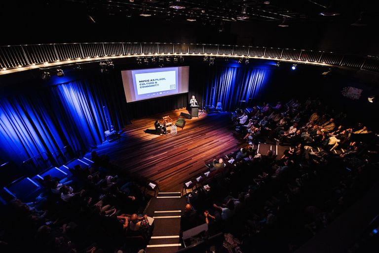 Wide overhead view of a speaker at the podium during the Remote and Regional Summit 2025 presentation in a theater.